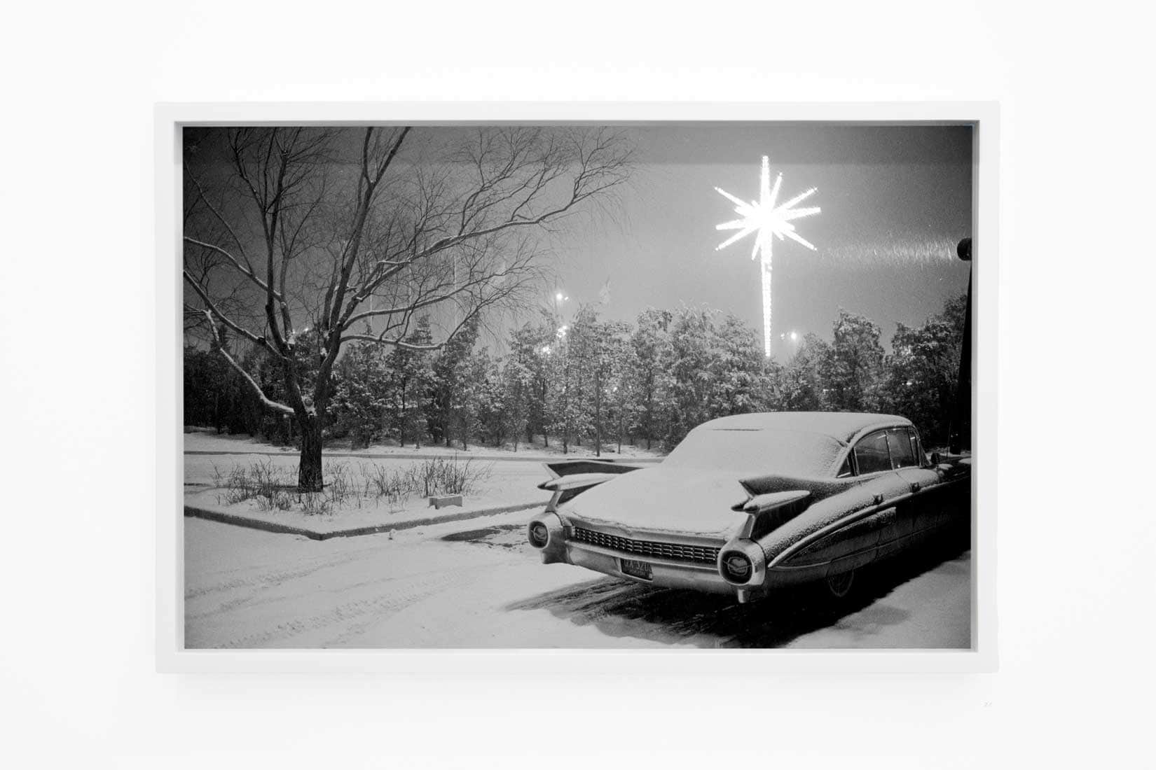 Black and white photo of a car in snow and fireworks in the sky.